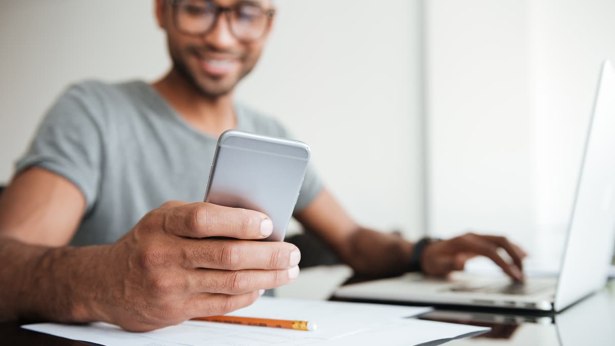 Jüngerer Mann sitzt auf Schreibtisch vor Laptop und schaut auf sein Smartphone in der Hand